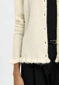Off-white fuzzy cardigan with small buttons and ruffled hem worn over black pants with a black leather belt featuring a gold buckle.