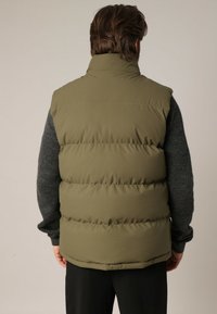Olijfgroen puffer vest met een hoge kraag, voorzien van horizontale stiksels en een gladde afwerking, gedragen over een donkergrijze sweater.