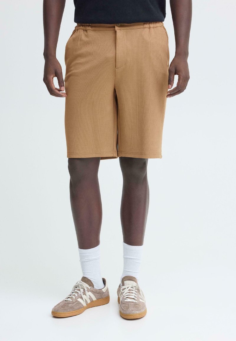 Shorts marron jusqu'aux genoux en tissu texturé avec une ceinture élastique ; portés avec des chaussettes blanches et des baskets beiges.