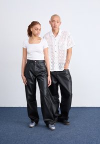 Twee personen staan naast elkaar, de een in een nauwsluitend wit T-shirt en zwarte leren broek, de ander in een wit geperforeerd overhemd met knopen en zwarte broek.