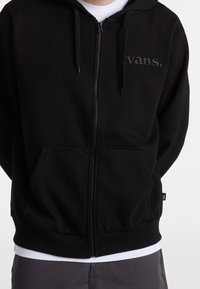 Person trägt einen schwarzen Vans-Kapuzenpullover mit Reißverschluss über einem weißen Hemd, graue Hose, die Hände hinter dem Rücken auf einem einfarbigen Hintergrund.