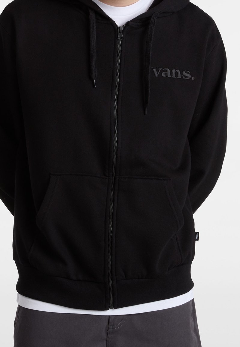 Person trägt einen schwarzen Vans-Kapuzenpullover mit Reißverschluss über einem weißen Hemd, graue Hose, die Hände hinter dem Rücken auf einem einfarbigen Hintergrund.