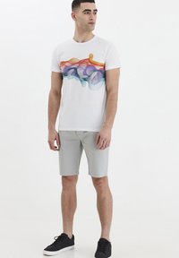 Camiseta blanca con un diseño abstracto colorido en naranja, azul y púrpura. Combinada con pantalones cortos gris claro y calzado negro.