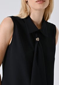 Donna bionda che indossa una blusa nera senza maniche con un ampio colletto, dettaglio drappeggiato sul davanti tenuto da una chiusura dorata e orecchini a pendente dorati.