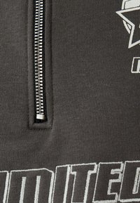 Sudadera con cremallera gris y cremallera plateada, de tejido texturizado. Presenta letras en negrita en blanco a lo largo de la parte inferior y un gráfico de estrella encima.