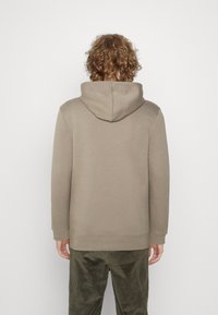 Bluza z kapturem w jasnobrązowej tkaninie, o luźnym kroju, z prążkowanymi mankietami i obszyciem. Noszona z oliwkowozielonymi spodniami sztruksowymi.