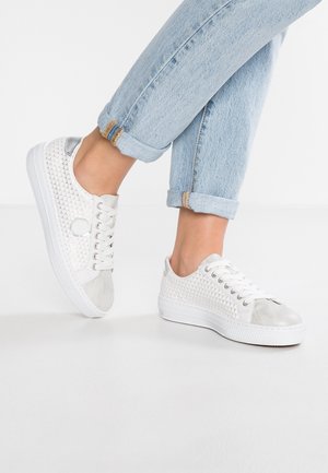 Pieds portant des baskets blanches basses avec un motif hexagonal et des jeans clair retroussés sur un fond blanc.