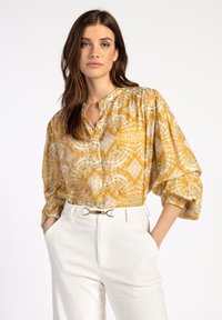 Gelbe gemusterte Bluse mit Stehkragen, Knopfverschluss und luftigen Ärmeln. Kombiniert mit weißen hochgeschnittenen Hosen, die einen goldenen Gürtel haben.