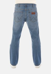 Vista trasera de una persona que lleva pantalones vaqueros de mezclilla azul claro con una "W" bordada en los bolsillos traseros y zapatillas blancas sobre un fondo liso.