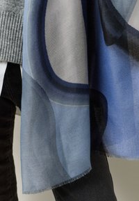 Blue and gray patterned scarf made of lightweight fabric, featuring soft fringing along the edges, draping elegantly over a gray sweater.