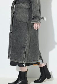 Osoba v vybledlém černém džínovém kabátu s velkou kapsou, vzorované midi sukni a černých kotníkových botách na bílém pozadí.