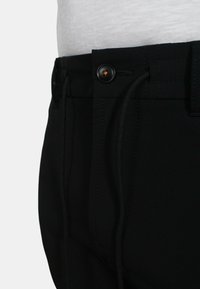 Pantaloni neri con vita regolabile tramite cordino, chiusura a bottone e texture del tessuto liscia. Il design include dettagli di cucitura sottili.