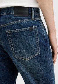 Jeans de mezclilla azul oscuro con un bolsillo trasero visible y una etiqueta de cuero en la cintura. Presentan costuras en contraste y una textura suave.