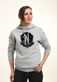 Donna con capelli castani in felpa grigia con logo scudo in bianco e nero, jeans neri, in piedi con una mano sulla vita, sfondo neutro.