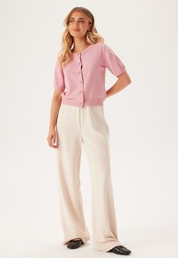 Kortärmad rosa cardigan i strukturerad tyg med knappstängning, tillsammans med krämfärgade vida byxor och svarta slip-on skor.