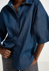 Denim skjorte med knaplukning, afslappet pasform, korte ærmer, mørkeblå farve og synlig sidesøm. Standard knap-lukning detalje.