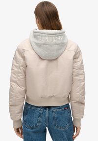 Lyserød bomberjakke med en grå hætte, har ribbede manchetter og kant, samt Superdry-logo broderet på hætten.