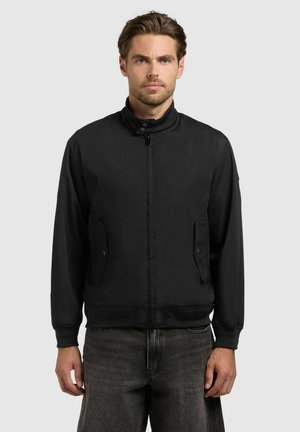 Mężczyzna o jasnobrązowych włosach, ubrany w czarną kurtkę zapinaną na zamek i ciemnoszare dżinsy, stojący przodem na jednolitym tle.