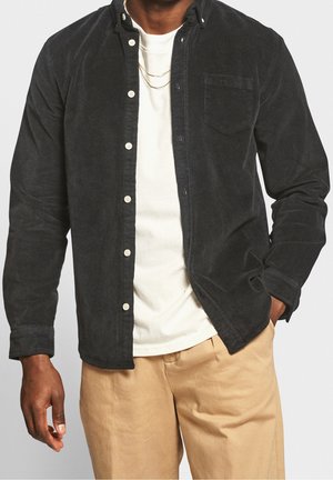 Homme portant une chemise en velours côtelé noire boutonnée sur un t-shirt blanc, un pantalon beige, et deux fines chaînes en or, avec une main dans la poche.