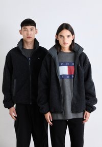 Chaquetas de forro polar negras con cuellos altos, cierres de cremallera y puños elásticos, que se llevan sobre camisetas grises con un diseño de logotipo geométrico.