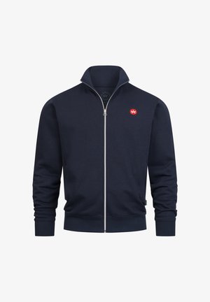 Laivastonsininen vetoketjullinen takki korkealla kauluksella, valmistettu pehmeästä kankaasta. Vasemmassa rinnassa pieni punainen logo, resorit hihansuissa ja helmassa.