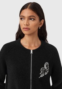 Cardigan nero con zip frontale, tessuto strutturato, maniche raglan e un design ricamato sul lato sinistro del petto con dettagli intricati.