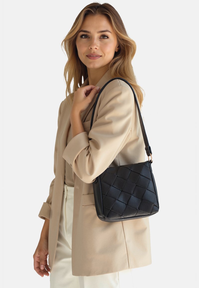 Schwarze Handtasche mit gewebter Textur, flachem Boden und einem einzelnen Schultergurt, getragen von einer Person in einem beigen Blazer.