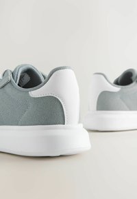 Lysegrå sneakers med en ruskindsoverflade og hvide gummisulder. Har en glat hvid hæl og syninger som accenter. Rund tå design.
