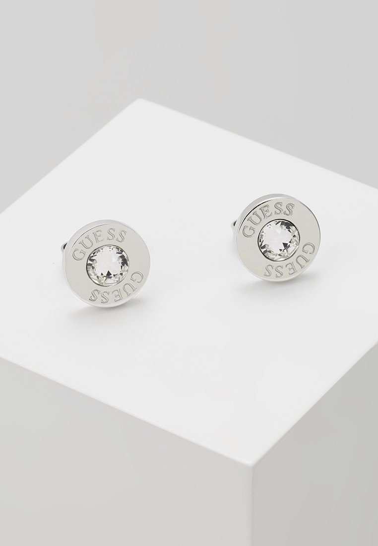 Boucles d’oreilles puces rondes en argent avec un centre en cristal clair et le nom de la marque Guess gravé autour du bord.