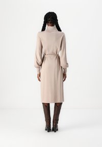 Vestito lungo beige con collo alto, vita stretta e maniche a sbuffo, abbinato a stivali marroni al ginocchio. Tessuto liscio.