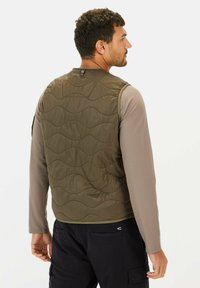 Leichte, olivgrüne, gesteppte Weste mit wellenförmiger Textur, hohem Kragen und Druckknopfverschluss hinten, getragen über einem langärmeligen beige Oberteil.