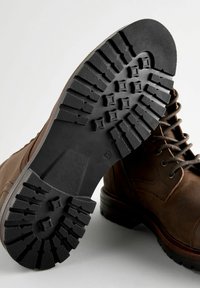 Bota de cuero marrón con suela de goma negra. La suela presenta un patrón de textura para mayor agarre, y la parte superior tiene cordones y ojales.