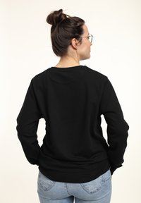 Donna con capelli scuri raccolti in un bun, che indossa occhiali, una maglietta nera a maniche lunghe e jeans azzurri chiari, in piedi con le mani sui fianchi, voltata di spalle.
