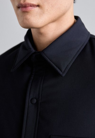 Camicia con colletto rigido, realizzata in tessuto scuro e liscio. Presenta una placca frontale con un bottone e una tasca sul petto.