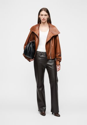 Kobieta ubrana w brązową skórzaną kurtkę, biały top bez rękawów, czarne skórzane spodnie, brązowe szpiczaste szpilki, trzymająca dużą czarną torebkę.