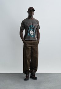 Άνδρας που στέκεται με τα χέρια στις τσέπες, φοράει καφέ καπέλο, σκούρο γραφιστικό μπλουζάκι με σχέδιο φιδιού και κρανίου, χαλαρά καφέ παντελόνια και σκούρα μποτάκια.