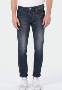 Man in dark wash slim-fit opgerolde jeans en witte sneakers, staande tegen een effen lichte achtergrond.