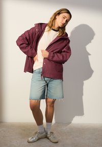 Vínovo červená nepremokavá bunda s kapucňou, elastickými manžetami a prednými vreckami, v kombinácii so svetlomodrými džínsovými šortkami a striebornými teniskami.