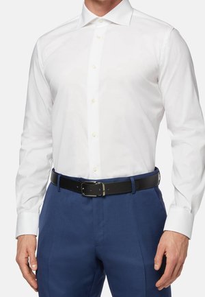 Mężczyzna w białej, długorękawowej koszuli zapinanej na guziki, włożonej do niebieskich spodni z czarnym paskiem z srebrną klamrą.