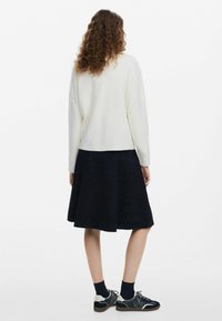 Suéter blanco de canalé, falda de mezclilla azul marino acampanada con patrón texturizado, zapatillas negras con detalles y calcetines hasta el tobillo. Vista desde atrás.
