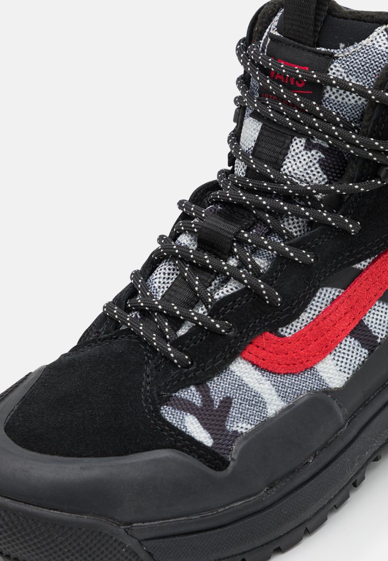 Zapatilla alta con ante negro y textil estampado en gris y blanco. Presenta un logo rojo característico y suela de goma negra texturizada.