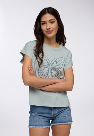 T-shirt in cotone azzurro chiaro con maniche corte, adornato con un design di farfalla in accenti argentati. Abbinato a pantaloni shorts di jeans.