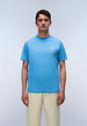Mann blickt nach vorne, trägt ein schlichtes blaues T-Shirt und eine hellbeige Hose vor hellem, einfarbigem Hintergrund.