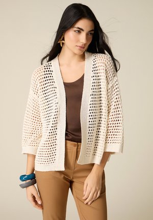 Kvinde iført beige åbenstrikket cardigan over brun top og beige bukser, accessoriseret med søstjerne-øreringe og masser af armbånd, mod en neutral baggrund.