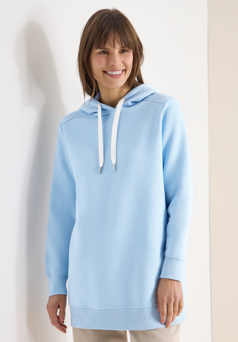 Femme souriante aux cheveux bruns portant un sweat à capuche bleu clair avec des cordons blancs, debout devant des murs blancs et beiges.