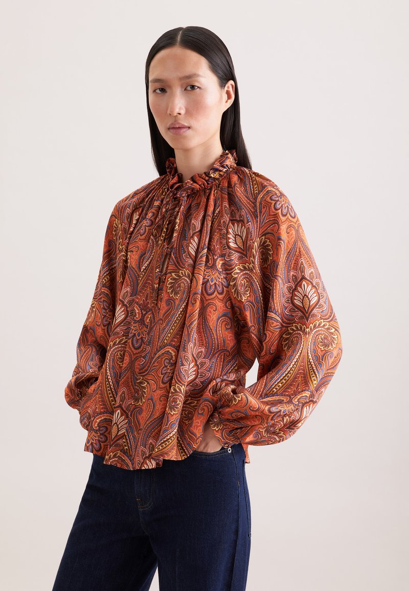 Femme aux longs cheveux noirs portant un chemisier ample à manches longues avec un motif cachemire orange et un jean bleu foncé, les mains dans les poches.
