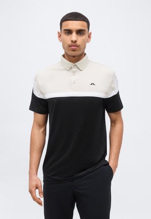 Νέος άνδρας με κοντά σκούρα μαλλιά που φοράει κοντομάνικο πόλο πουκάμισο δύο τόνων με μπεζ πάνω μέρος, μαύρο κάτω μέρος και λευκή λωρίδα στο στήθος.