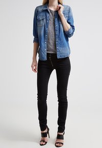 Denim button-up shirt in light blue, featuring two chest pockets, worn over a gray top, paired with black skinny jeans and black heeled sandals.