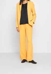 Gul skräddarsydd kostym med en svart blus, som har en avslappnad passform och långa byxor. Slätt tyg med en subtil lyster. Svarta skor fulländar outfiten.