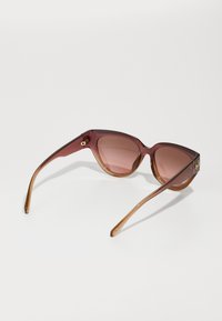 Óculos de sol de tom castanho com forma geométrica de olhos de gato, armações transparentes e hastes castanhas claras com detalhes em dourado nas dobradiças.